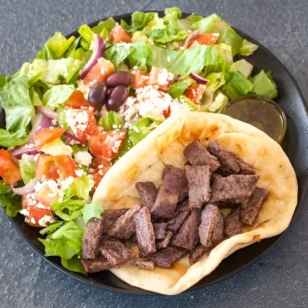 Mediterranean gyro plate with sliced meat on pita bread, Greek salad, and a side of dressing.