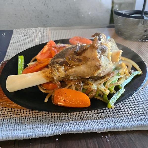 A steaming lamb shank with bone, bell peppers, onions, and tomatoes on a black plate.
