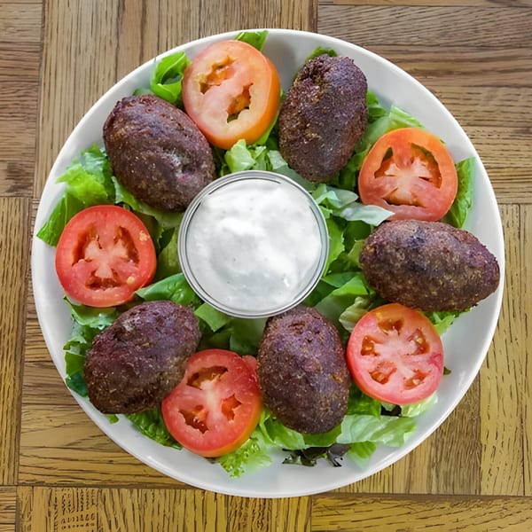 Kibbeh, sliced tomatoes, and lettuce with a side of white sauce on a white plate.