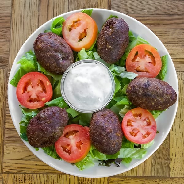 Kibbeh, sliced tomatoes, and lettuce arranged around a white dipping sauce on a white plate.