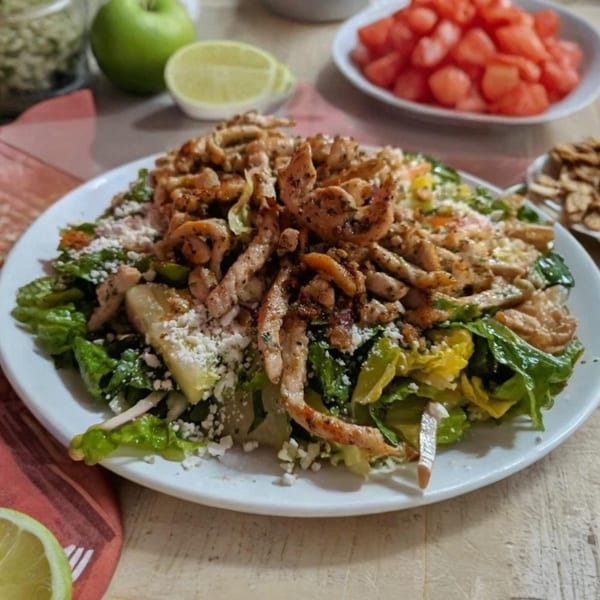 A Mediterranean chicken salad with lettuce, feta, and apples, with lime and watermelon in the background.