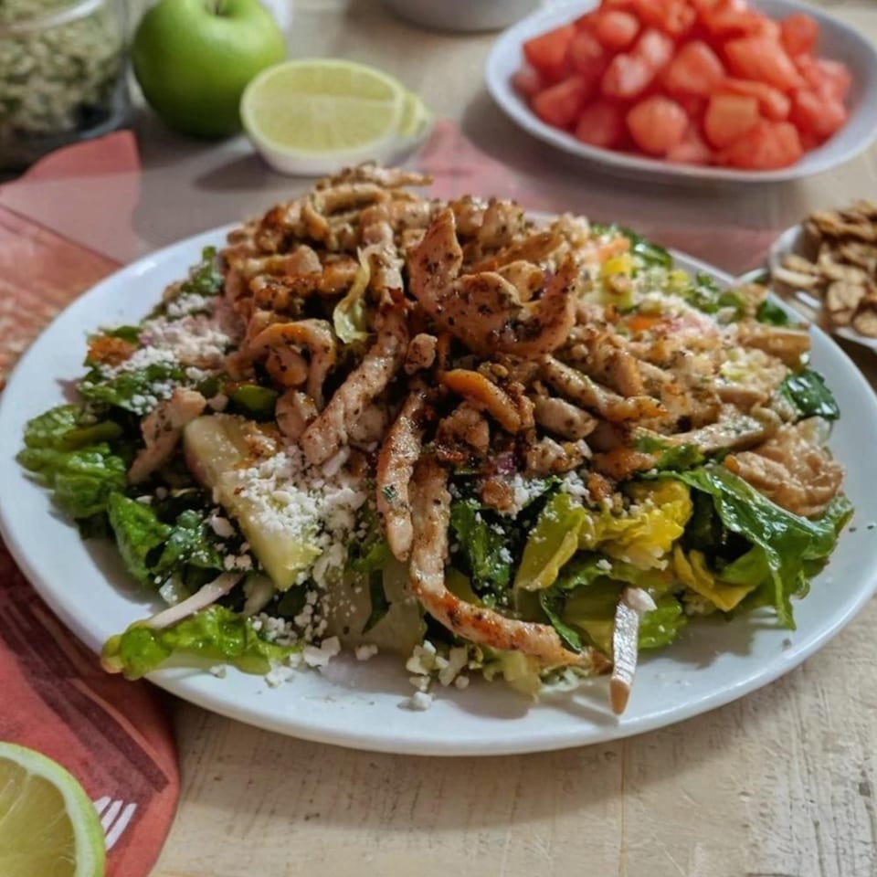 A Mediterranean chicken salad with lettuce, feta, and apples, with lime and watermelon in the background.