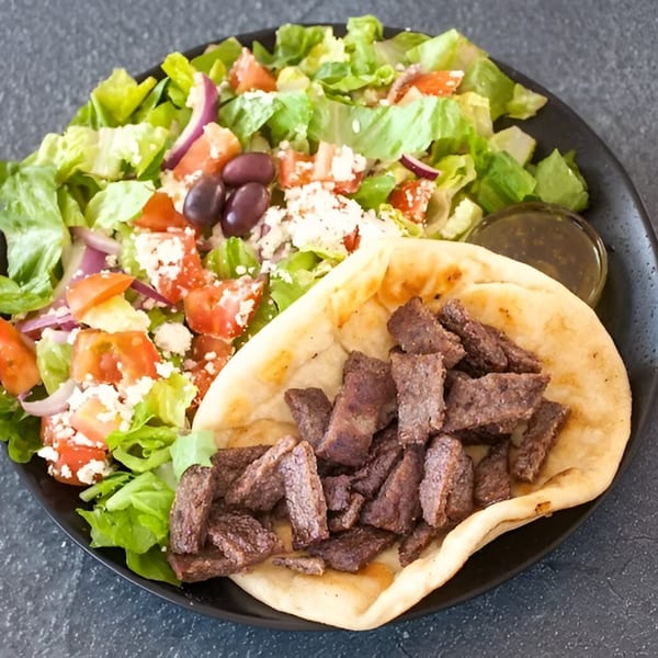 A Mediterranean gyro plate with sliced meat on pita bread, a Greek salad, and a side of sauce.