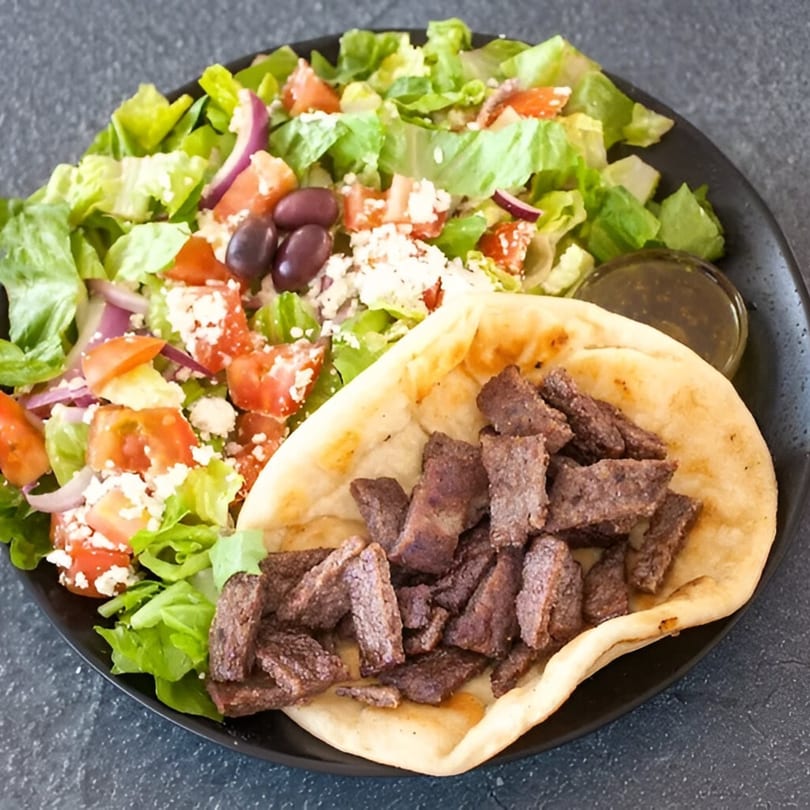 A Mediterranean gyro plate with sliced meat on pita bread, a Greek salad, and a side of sauce.